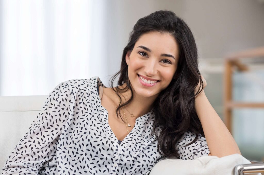 Smiling woman with clear, healthy-looking skin relaxing indoors after aesthetic wellness treatment.
