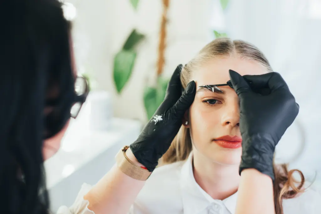 Close-up of a beauty technician wearing black gloves shaping or tinting a woman’s eyebrows with a small brush, as the client looks forward in a salon setting.