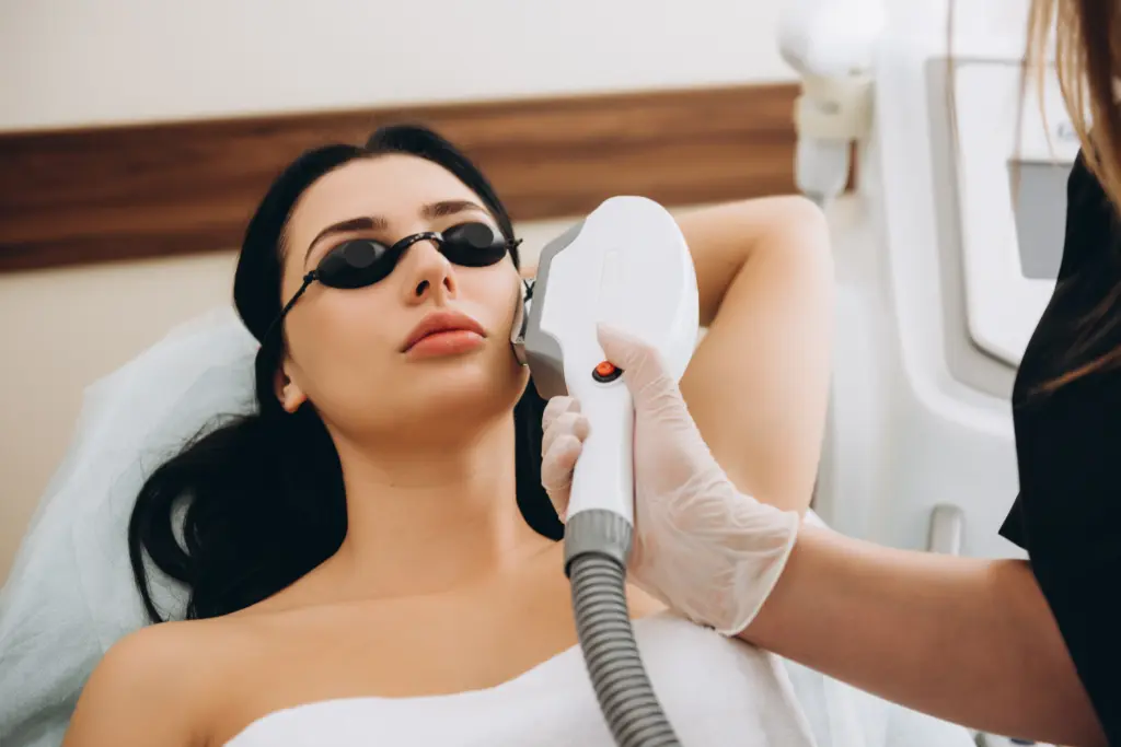 Woman wearing protective goggles receiving a facial laser treatment on her cheek with a handheld device in a medical spa setting.